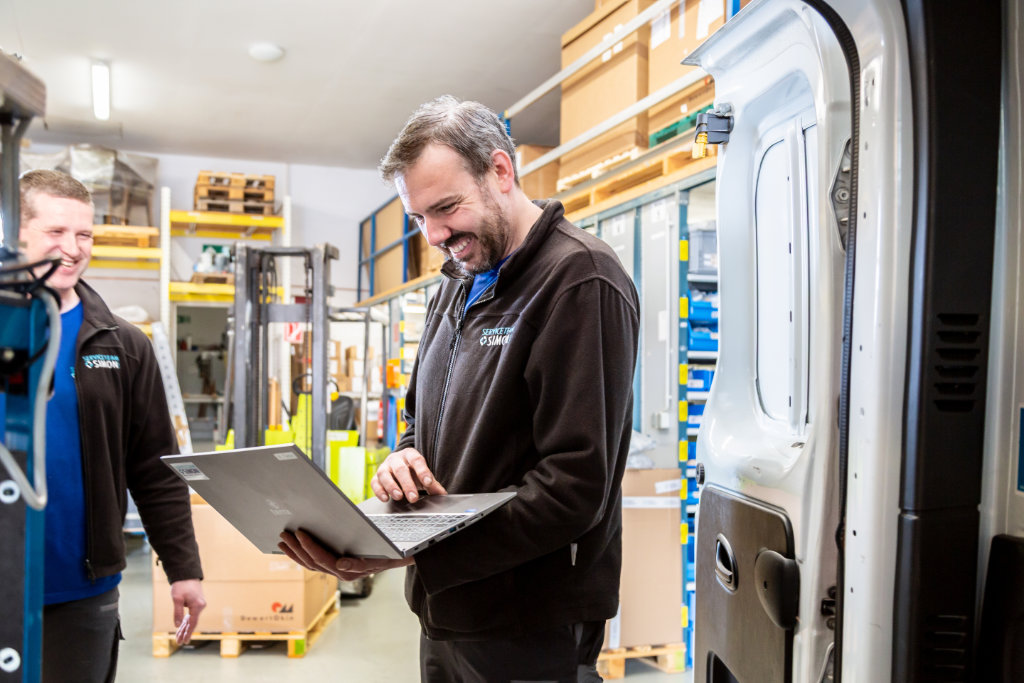 Ein freundlicher Mitarbeiter vom Serviceteam Simon steht im Lager und schaut mit einem Kollegen auf einen Laptop.