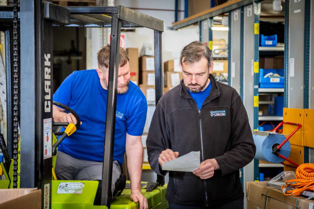 Ein Mitarbeiter vom Serviceteam Simon sitzt im Gabelstapler und schaut mit einem Kollegen auf ein Blatt Papier. Beide tragen Firmenkleidung.