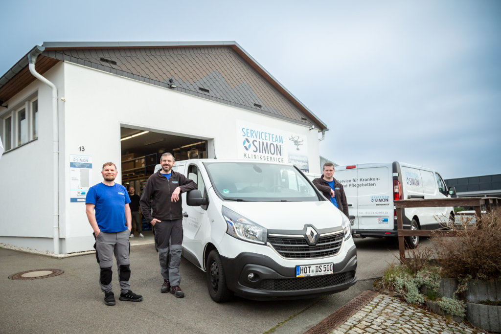 Drei Mitarbeiter mit blauen Shirts, neben einem weißem Lieferwagen und vor dem Gebäude von SERVICETEAM SIMON KLINIKSERVICE.