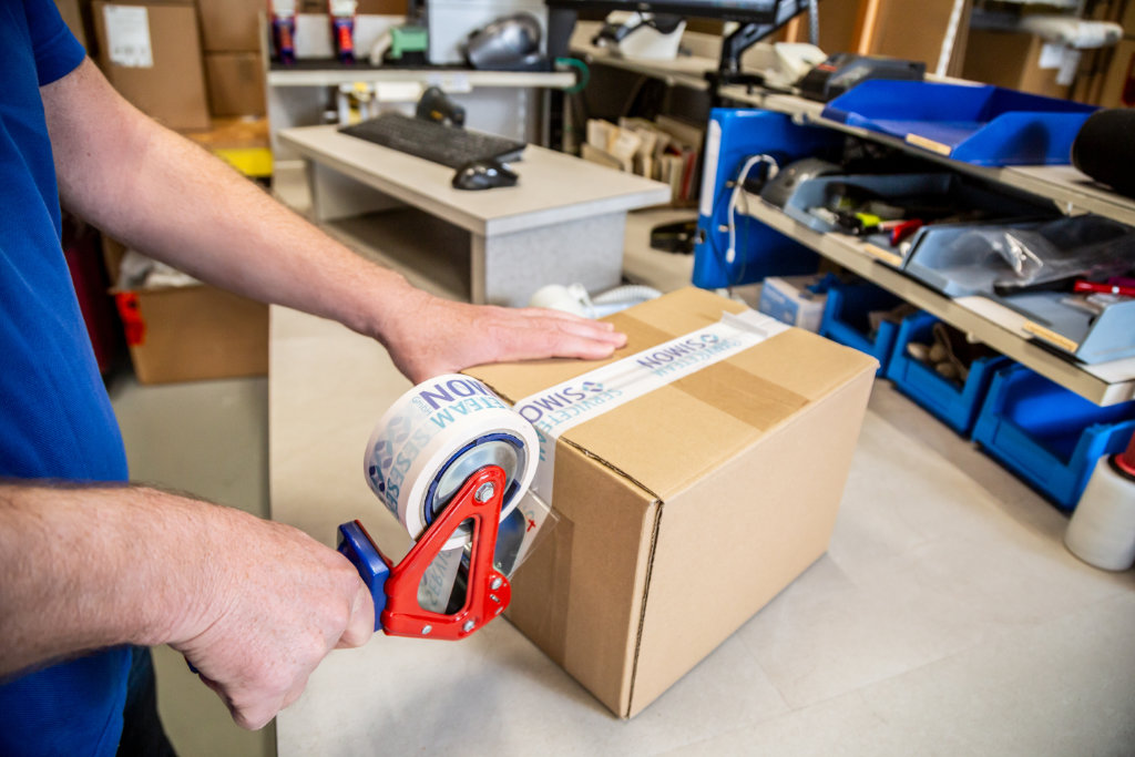 Ein Mitarbeiter vom Serviceteam Simon verpackt ein Karton mit Logo-Klebeband – als Vertriebspartner im Bereich Medizintechnik und Logistik.