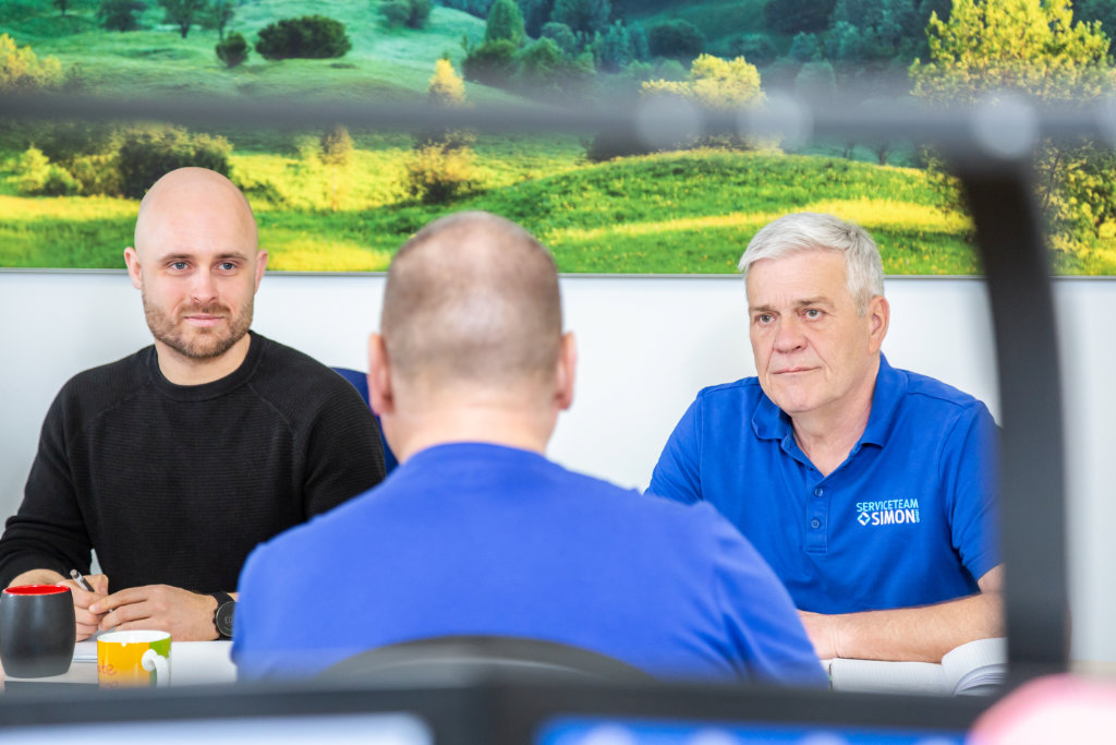 Das Service Team Simon sitzt in einer Besprechungsrunde, die Mike Richter und Dieter Simon in einer Frontalansicht sowie einen weiteren Mitarbeiter verschwommen von hinten zeigt, in einem hellen freundlichen Raum, an dessen Wand ein Bild mit einem Naturmotiv hängt.