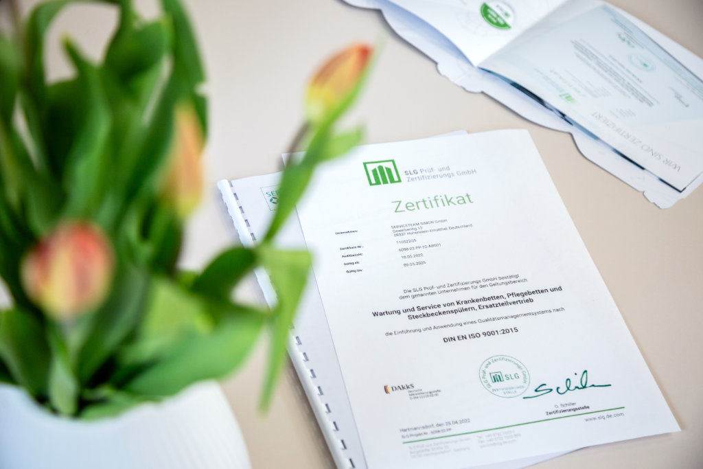 Blumen im Vordergrund dahinter auf dem Tisch liegt ein ISO 9001:2015-Zertifikat für das Serviceteam Simon – Qualitätsmanagement für Medizintechnik.