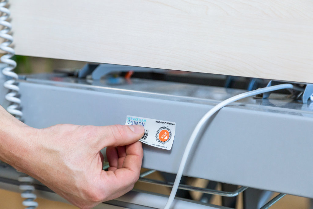 Ein Mitarbeiter vom Serviceteam Simon klebt ein Prüfetikett mit dem Hinweis ‚Nächster Prüftermin‘ an einem medizinischen Gerät – Teil der technischen Analyse und Wartung.