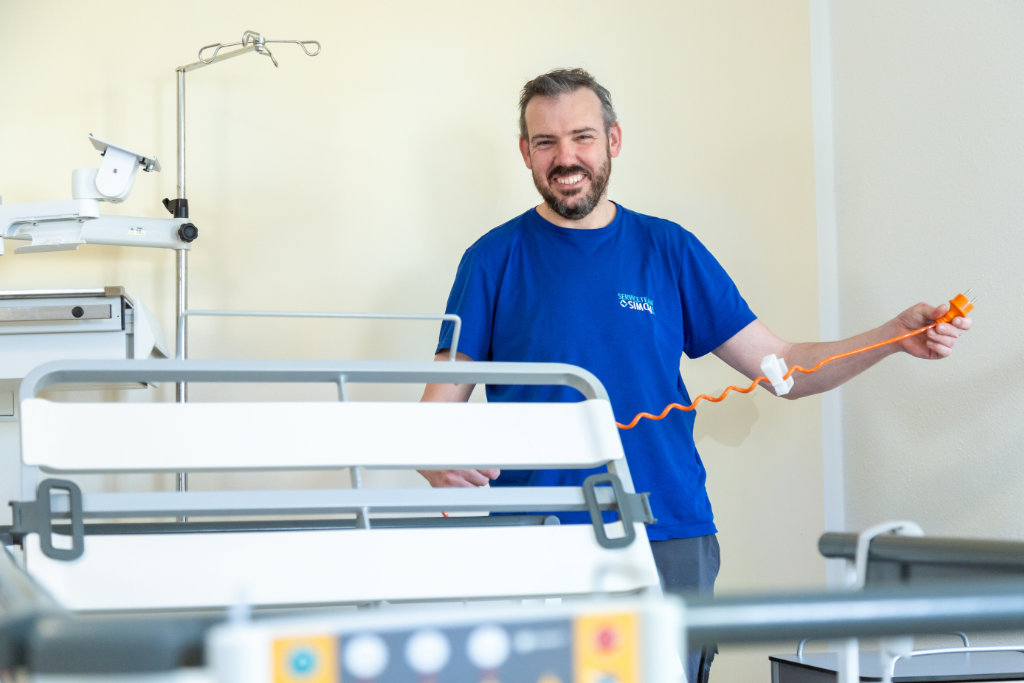 Ein freundlicher Mitarbeiter vom Serviceteam Simon steht hinter einem höhenverstellbaren Krankenbett und hält einen orangefarbenen Rufknopf in der Hand. Er lächelt direkt in die Kamera.